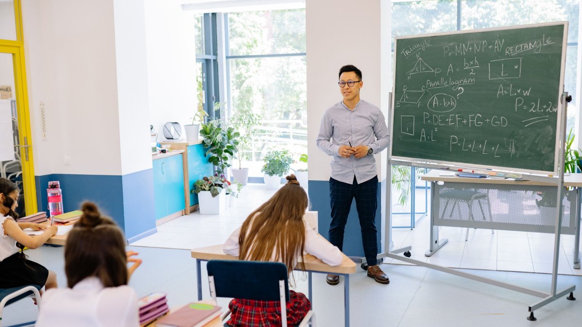 Professor de pé diante do quadro com fórmulas de geometria, explicando a matéria para alunas sentadas em suas carteiras